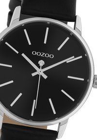 Montre noire avec un boîtier circulaire en argent, design minimaliste, marqueurs d'heure argentés et un bracelet en cuir noir. Caractérise le nom de la marque "OOZOO."
