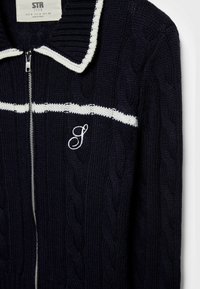 Pull en maille torsadée bleu marine avec fermeture éclair, bande blanche traversant la poitrine, bordure blanche au col et lettre "S" brodée sur le devant gauche.