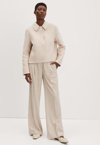 Beige wollen cropped jacket met een kraag, gecombineerd met crèmekleurige wijde broekspijpen. Het model draagt beige instap schoenen met geplooide details.