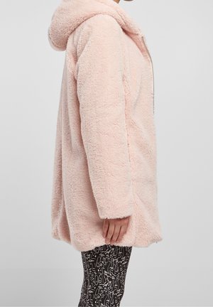Mujer vistiendo una suave chaqueta de forro polar con capucha de color rosa claro y cremallera, combinada con leggings de patrón abstracto en blanco y negro.