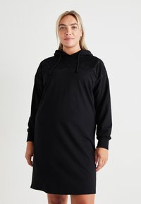 Črna obleka s kapuco iz bombaža, ki ima čipkaste detajle na ramenskem delu in sproščen fit. Dolgi rokavi in sprednji kenguru žep.