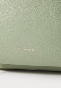 Zöld, texturált bőr kézitáska arany "Coccinelle" logóval, sima felület és strukturált forma jellemzi.