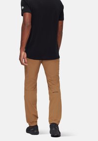 Bruine outdoorshorts met een getailleerd ontwerp, gebogen knieën en een zwart logoaccent op de kuit. G gedragen met een zwart T-shirt met korte mouwen.
