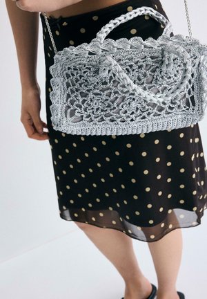 Donna con gonna nera a pois che tiene una borsa all'uncinetto grigio chiaro con tracolla a catena su sfondo neutro.