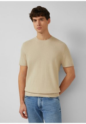 Młody mężczyzna o krótkich kręconych włosach, ubrany w beżowy, teksturowany, krótki sweter z krótkim rękawem i jasnoniebieskie dżinsy, na tle jednolitego szarego tła.