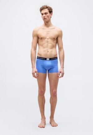 Jonge man staat op blote voeten, draagt blauwe boxer briefs met een zwarte tailleband waarop "Polo Ralph Lauren" staat, tegen een effen grijze achtergrond.