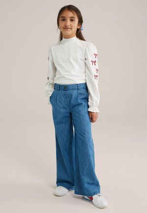 Maglietta bianca a maniche lunghe con colletto arricciato e ricamo di un fiocco rosso sulle maniche, abbinata a pantaloni larghi in denim e sneakers bianche.