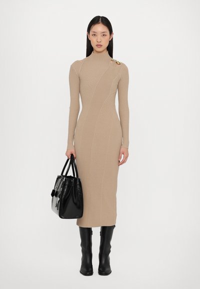 Beige ribbed long-sleeve dress with high neck and side accent detail. Model holds a black leather handbag. Knee-high black boots.