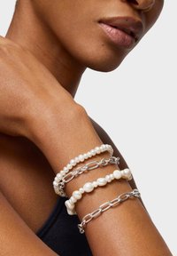 Zbliżenie na nadgarstek noszący dwie bransoletki z pereł oraz dwie srebrne bransoletki z ogniw łańcucha na gładkiej, brązowej skórze.