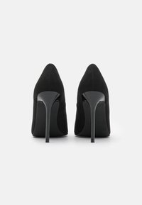 Černé vysoké podpatky s špičatou špičkou, vyrobené z měkké semišové látky, s lesklými černými stiletto podpatky a hladkou podrážkou.