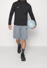 Felpa nera con cappuccio, pantaloni sportivi grigi, calze nere con motivo e scarpe da ginnastica nere. Tiene in mano un pallone da basket azzurro.