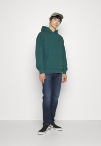 Sudadera de algodón verde con bolsillo tipo canguro, combinada con jeans azul oscuro y zapatillas negras. Una gorra caqui completa el atuendo.