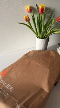 Brun papirpose med krøllet tekstur, prydet med et trykt logo i hvidt og orange. Grønne blade og rød-gule tulipaner i en ribbet hvid vase i nærheden.
