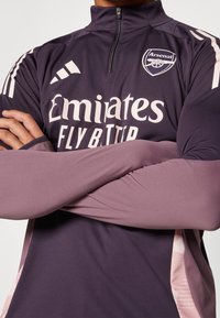 Lila Trikot mit hochgeschlossenem Kragen und Reißverschluss, ausgestattet mit dem weißen „Emirates“-Logo und dem „Arsenal“-Emblem. Lange Ärmeln mit strukturierten Akzenten.