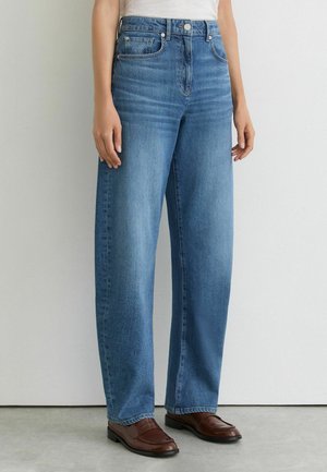 Kvinde iført højtaljede blå straight-leg jeans med forlommer, hvid top og brune læderloafers, stående mod en lys væg.