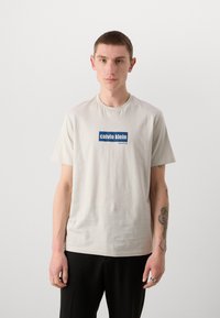 Lys beige bomulls T-skjorte med en blå rektangulær logo med teksten "calvin klein" og "new york city", rund hals, korte ermer.