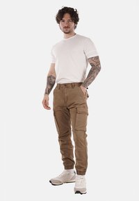 Pantaloni cargo beige con molte tasche, polsini elasticizzati e una texture liscia, abbinati a una t-shirt bianca e sneakers chiare.