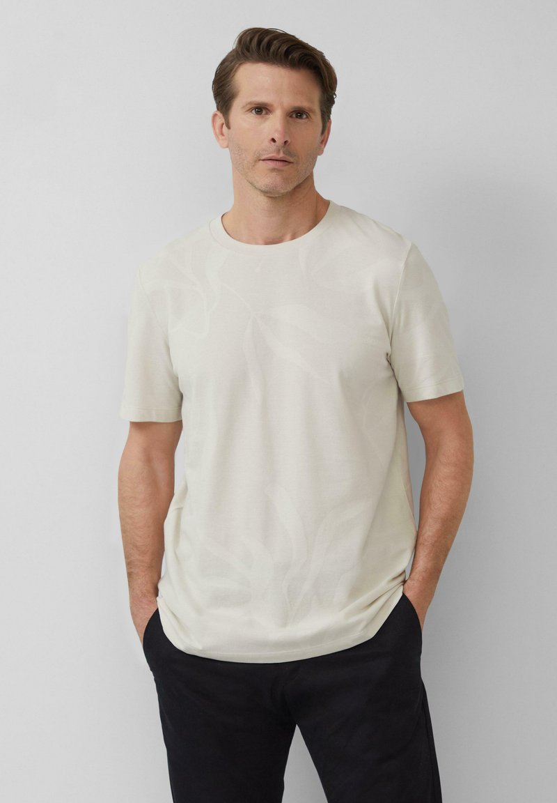 Helles beiges T-Shirt mit kurzen Ärmeln, das ein dezentes Blattmuster aufweist. Hergestellt aus weichem Stoff, entworfen mit einem runden Ausschnitt und lockerer Passform.