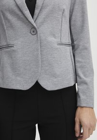 Šedý blazer s hladkou texturou, zapínání na jeden knoflík a dvě vertikální kapsy. Přiléhavý design, kombinovaný s černými kalhotami.