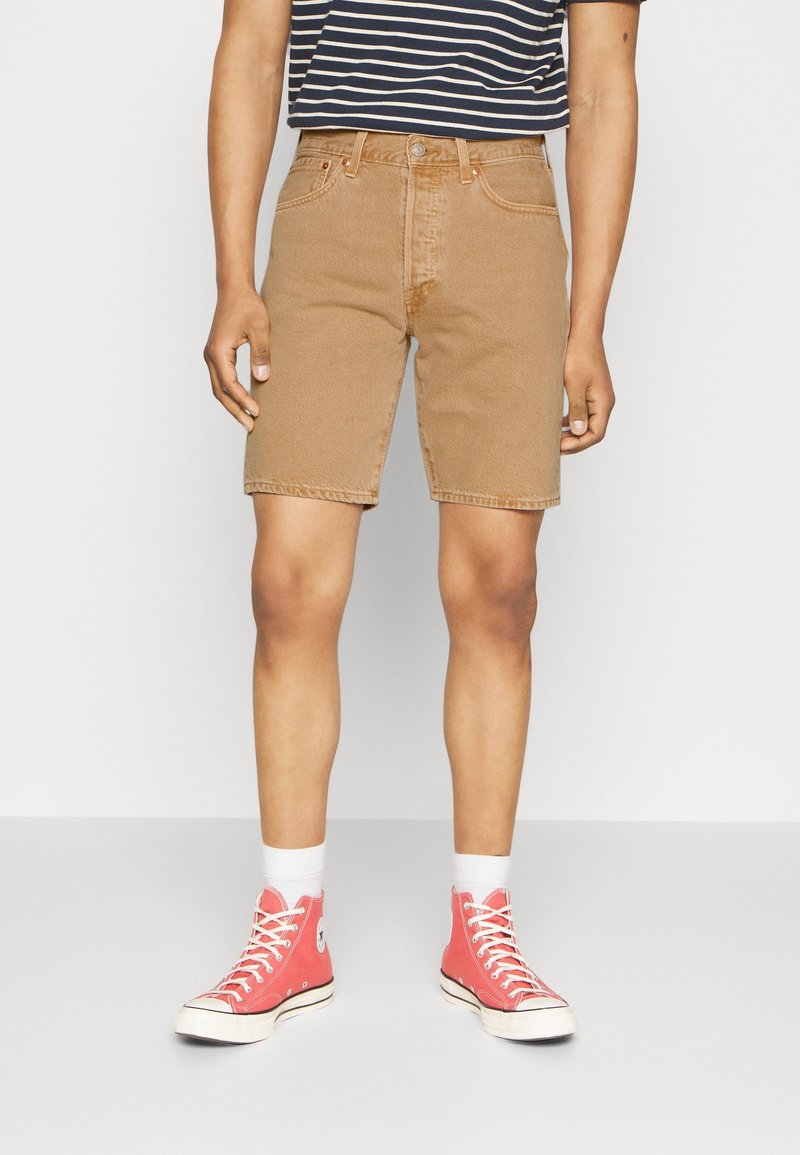 Shorts en denim marron à coupe droite, avec poches avant et ourlet cousu. Associés à des baskets en toile rouges et des chaussettes blanches.