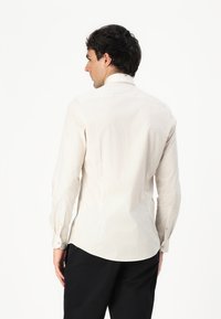 Cămașă bej deschis, cu nasturi, cu un model subtil, mâneci lungi, spate cu yoke și hem ondulat. Țesătura pare netedă și ușor texturată.