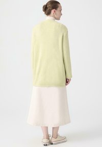 Vrouw die met haar rug naar de kijker staat, gekleed in een lichtgroene ribbeltrui, een beige midi-rok, beige sneakers en gouden oorbellen, met haar in een bun.