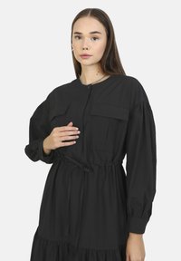 Robe noire à manches longues avec fermeture à boutons, deux poches poitrine, taille élastique et jupe à volants. Tissu doux et lisse avec un léger éclat.