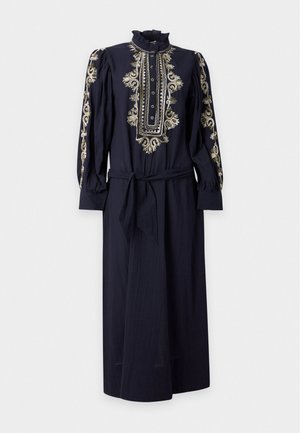 Femme debout portant une longue robe bleu marine avec broderies beiges sur les manches et le corsage, taille nouée, et chaussures plates beiges sur fond blanc.