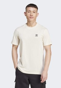 Krémové bavlněné tričko s krátkými rukávy, kulatým výstřihem a malým černým logem Adidas na levé části hrudi. Design v uvolněném střihu.