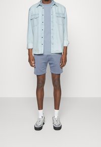 Camicia di jeans azzurro chiaro, t-shirt blu, pantaloni corti coordinati e sneakers grigie con suola nera. Dettagli notevoli includono tasche con bottoni.