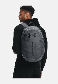 Zaino grigio dal design elegante, con il logo Under Armour e tessuto testurizzato. Include spallacci regolabili e una tasca frontale con zip.