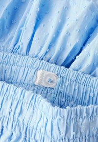 Tela azul claro con textura de puntos elevados, que presenta una cintura con frunces y un detalle de botón. Material suave y ligero.