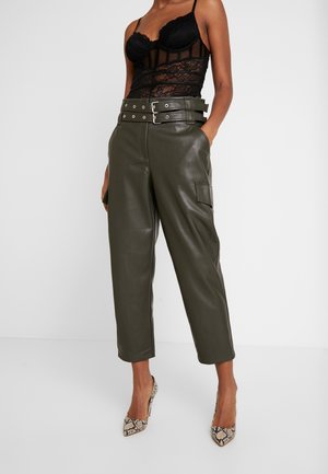 Frau trägt hoch taillierte, olivgrüne Lederhosen mit doppeltem Schnallengürtel, schwarzes, transparentes Spitzenoberteil und Schlangenleder-High Heels, steht mit den Händen in den Taschen.