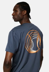Marineblå t-shirt med kort ärm i mjukt tyg, med en rund orange och vit grafisk design på baksidan.