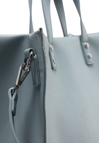 Jasnoniebieska skórzana torebka o teksturowanej powierzchni, z srebrnymi akcentami i długimi uchwytami. Posiada zamek błyskawiczny i widoczne detale ściegów.