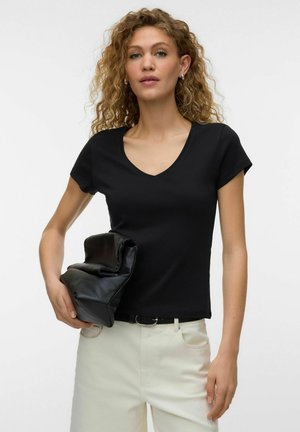 Kvinde med krøllet hår iført sort T-shirt med V-hals og hvide bukser, der holder en foldet sort lædertaske mod en hvid baggrund.