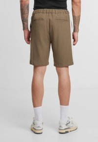 Shorts marron avec une ceinture élastique, deux poches arrière et des bandes blanches sur les côtés. Confectionnés dans un tissu lisse, longueur au niveau des genoux.