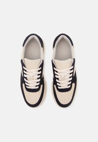 Beige och marinblå sneakers med en texturerad mocka utsida, perforerade detaljer på tån och platta krämfärgade skosnören. Minimalistisk design.