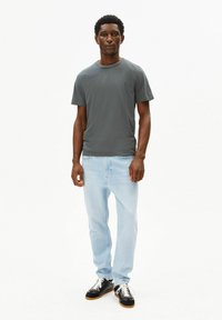 Graues T-Shirt mit kurzen Ärmeln, hellblaue Jeans und schwarze Sneakers mit weißen Schnürsenkeln. Das Modell steht aufrecht vor einem schlichten weißen Hintergrund.