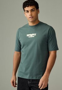 Tricou din bumbac de culoare teal închis, cu mâneci scurte și gât rotund. Prezintă un design grafic al textului în alb pe piept.