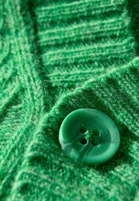 Grön stickad kavaj med ett texturerat mönster och en glansig, grön, fyrhålsknapp. Tyget ser mjukt och luddigt ut.