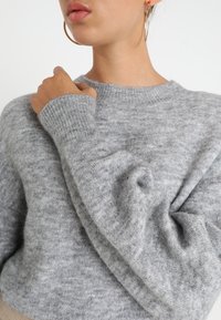 Ljusgrå tröja i mjukt, texturerat tyg, med rund halsringning och lösa ärmar. Virad tyg vid handleden.