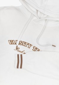 Hvid hoodie med en front kangaroo lomme, der har teksten "VARSITY TEAM" i brun. Brune striber på ærmekantene, blød stoftekstur.