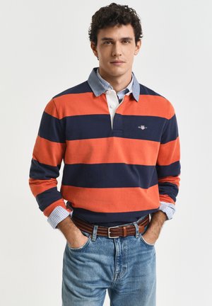 Mann trägt ein orange- und marineblau gestreiftes Rugbyhemd über einem blau karierten Hemd mit Knöpfen, Jeans mit braunem Gürtel, Hände in den Taschen, neutraler Hintergrund.