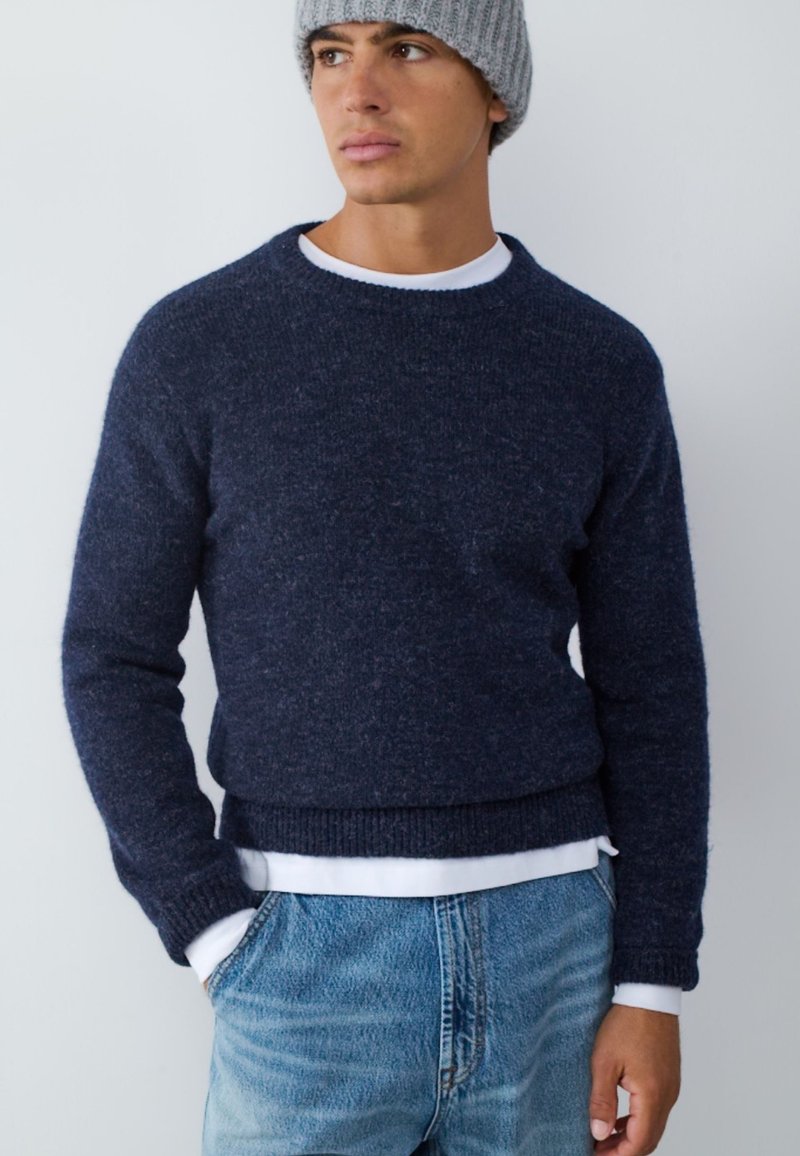 Jonge man draagt een donkerblauwe trui over een wit overhemd, lichtblauwe spijkerbroek en een grijze gebreide muts, met handen in de zakken.