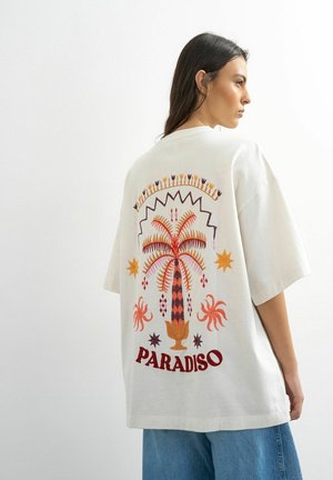 Femme aux longs cheveux noirs portant un t-shirt blanc oversize avec un palmier coloré et le texte « PARADISO » dans le dos, associé à un jean bleu.
