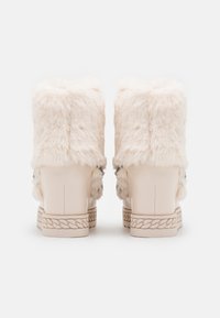 Botines de piel sintética en color crema pálido, con una suela beige lisa con diseño trenzado y herrajes decorativos en los laterales.