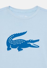 T-shirt di cotone azzurro chiaro con una grande grafica di coccodrillo blu e motivi triangolari, scollatura rotonda e maniche corte.