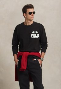 Jauns vīrietis valkā melnu Polo Motorsport džemperi, melnas bikses, apaļas saulesbrilles, ar sarkanu džemperi sasietu ap vidu, rokas kabatās.