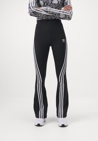 Svarta träningsleggings med vit tre-randsdesign längs sidorna. Åtsittande vid midjan, utsvängda vid anklarna, med en logotyp på höften.